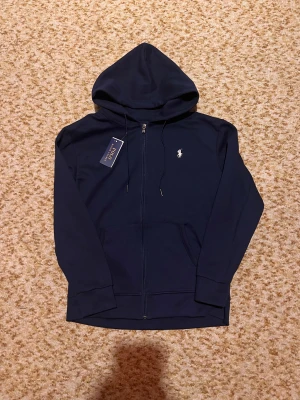 Marinblå hoodie från Polo Ralph Lauren - Snygg marinblå hoodie från Polo Ralph Lauren med vit broderad logga på bröstet. Tröjan har huva med snörning, dragkedja framtill och två praktiska fickor. Tillverkad i mjuk bomull och har långa ärmar. Perfekt för chill dagar och streetwear.