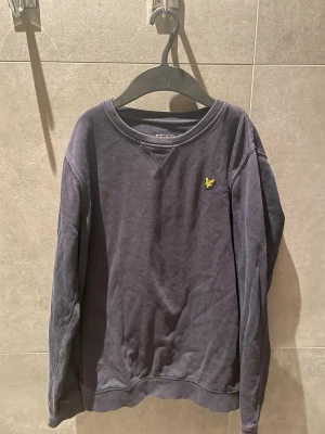 Mörkblå sweatshirt från Lyle & Scott - Mörkblå sweatshirt från Lyle & Scott med klassisk rund hals och lång ärm. Tröjan har det ikoniska gula örnlogot på bröstet och är tillverkad i mjuk bomull. Perfekt för dig som gillar stilrena och enkla plagg med snygg detalj.
