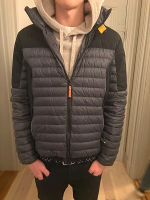 Parajumpers grå pufferjacka med röd huva - Säljer en snygg pufferjacka från Parajumpers i mörkgrått och svart med en fet röd huva. Jackan har quiltad design, lång ärm, dragkedja framtill och cool gul detalj vid kragen. Märkespatch på vänster ärm. Perfekt för dig som vill ha streetstyle med edge. Luvan går att stoppa in i en ficksa som man ser på sista bilden och det är en limiterad jacka. Hör av er vid frågor 