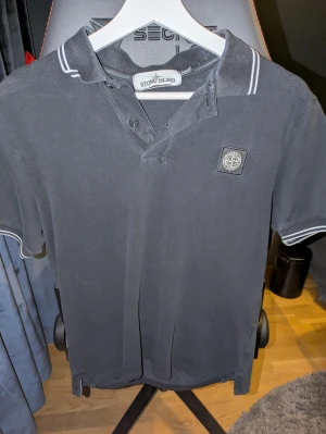 Svart pikétröja från Stone Island - Svart pikétröja från Stone Island med klassisk krage och vita ränder på krage och ärmslut. Tröjan har korta ärmar, knappar vid halsen och ikonisk Stone Island-logga på bröstet. Tillverkad i bomull och sitter slim på kroppen. i mycket bra skick.