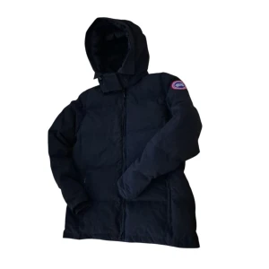 Svart dunjacka från Canada Goose macmillan  - Säljer en svart dunjacka från Canada Goose med huva och klassisk patch på ärmen. Jackan har dragkedja framtill och två sidofickor. Perfekt för kalla dagar och riktigt snygg till vinterlooken.