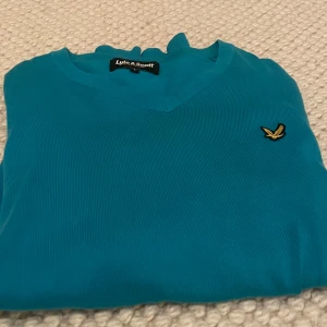 Turkos stickad tröja från Lyle & Scott - Snygg turkos stickad tröja från Lyle & Scott med v-ringning och klassisk gul logga på bröstet. Tröjan har långa ärmar och är gjord i mjukt material som känns skönt mot huden. Perfekt för dig som gillar stilrena och färgglada plagg.