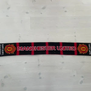 Manchester United halsduk svart/röd - Supporta ditt lag med denna klassiska Manchester United halsduk i svart och rött med klubbens logga och texten 'GLORY GLORY MAN UNITED' på sidorna. Halsduken har tydliga röda ränder och vita detaljer, perfekt för matchdagar och kalla dagar på läktaren.