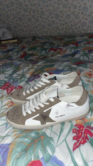 Golden Goose sneakers med stjärndetalj - Säljer ett par snygga Golden Goose sneakers i vit och beige med handskrivna detaljer och stjärnpatch på sidan. Skorna har rund tå, platt sula och klassisk snörning. Materialet är skinn och mocka, vilket ger en cool och lyxig vibe.