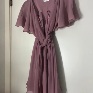 Ljuslila omlottklänning med volang - Superfin ljuslila omlottklänning med volangiga, vida ärmar och knyt i midjan. Klänningen har en flowy känsla och är gjord i ett tunt, lätt material som ger ett romantiskt intryck. Perfekt för sommarens alla tillfällen!