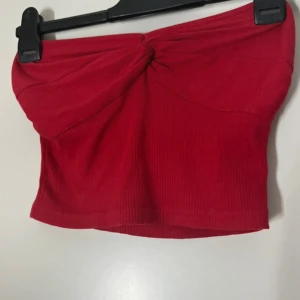 Röd ribbad tubtopp från Kappahl - Snygg röd tubtopp från Shein med ribbad struktur och twistad knut framtill. Toppen är bandeau-modell och sitter tight runt kroppen, perfekt för att skapa en trendig look. Enkel att matcha med både jeans och kjol.