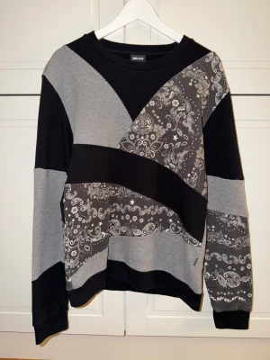 Svart och grå sweatshirt från Just Cavalli - Unik sweatshirt från Just Cavalli i svart och grått med coolt paisley-inspirerat mönster. Tröjan har långärmade paneler och kontrasterande block i olika nyanser av grått och svart. Perfekt för dig som gillar streetstyle och vill sticka ut.