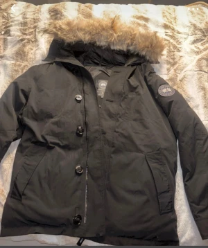 Svart dunjacka från Canada Goose - Säljer en svart dunjacka från Canada Goose med päls på huvan. Jackan har både dragkedja och knappar framtill, två stora fickor och klassisk patch på ärmen. Perfekt för kalla vinterdagar och har en snygg, tidlös look.