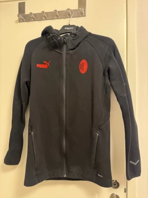Svart Puma hoodiejacka AC Milan M - Svart hoodiejacka från Puma med AC Milan-logga och röd Puma-text på bröstet. Jackan har huva med snörning, hel dragkedja och två fickor med dragkedja. Materialet är mjukt och sportigt, perfekt för dig som gillar fotboll och streetwear.