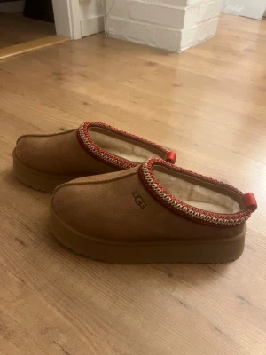 UGG W Tazz - Säljer ett par bruna slip-on skor från UGG med mjuk mocka på utsidan och fluffigt foder inuti. Skorna har en rund tå, platt sula och en snygg kant med röd och beige broderad detalj samt rött band bak. Perfekta nu till vintern och våren💕💕 skorna är helt oanvända och kartongen som skorna kom i finns med och de i den jag skickar dom i💕