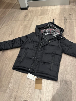 Svart pufferjacka från Burberry - Svart pufferjacka från Burberry med huva och rutig grå, vit och röd insida. Jackan har dragkedja och knappar framtill samt två sidofickor med dragkedja. Materialet är polyester och jackan har en loose passform som är perfekt för kalla dagar.