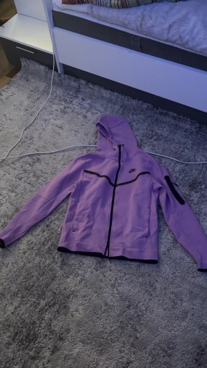 Lila Nike vindjacka med huva - Säljer en lila Nike vindjacka med svart dragkedja och detaljer. Jackan har huva, lång ärm och en extra ficka på ena ärmen. Perfekt för dig som vill ha en sportig och snygg look. Passar till både chill och aktivitet.