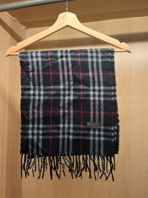 Burberry halsduk  - Snygg halsduk från Burberrys i 100% kashmir med klassiskt rutigt mönster i mörkblått, grått och röda linjer. Halsduken har fransar i ändarna och en mjuk, lyxig känsla. Perfekt för dig som vill ha en tidlös accessoar med premiumkänsla.