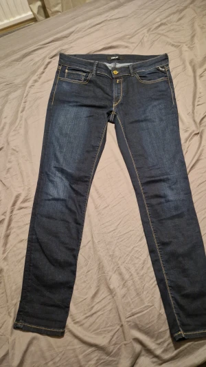 Replay jeans mörkblå  - Snygga Replay  jeans i mörkblå tvätt med klassiska kontrastsömmar och fem fickor. Modellen har slim passform och raka ben, med detaljer som Replay-logga på bakfickan och läderpatch i midjan. Perfekt för dig som gillar stilrena och moderna jeans.