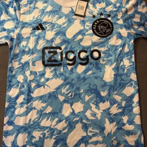 Ajax blåvit kortärmad fotbollströja - Ajax fotbollströja från Adidas i slim fit, storlek S. Tröjan har ett blått och vitt abstrakt mönster, svart Ajax-logga med tre stjärnor och Ziggo sponsortryck på bröstet. Tillverkad i lätt polyester, perfekt för match eller träning.
