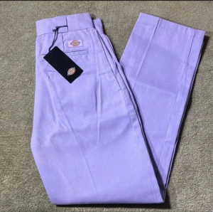 Dickies lila 574 W25/L30 - Säljer ett par ljuslila Dickies Phoenix Cropped byxor med raka ben och klassisk Dickies-logga ovanför bakfickan. Byxorna har croppad längd, bälteshällor och är tillverkade i slitstark bomull. Perfekt för dig som vill sticka ut med färg och stil.
