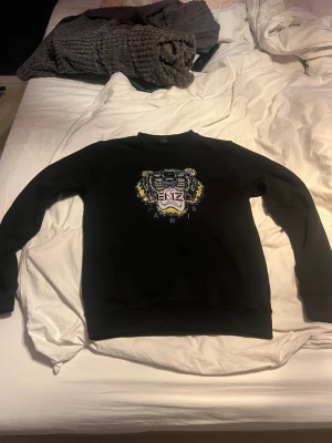 Svart Kenzo sweatshirt - Svart sweatshirt från Kenzo i storlek S med ikonisk broderad tiger på bröstet i blått, gult och beige. Tröjan har rund hals, långa ärmar och ribbade muddar. Perfekt för dig som vill ha en statement piece