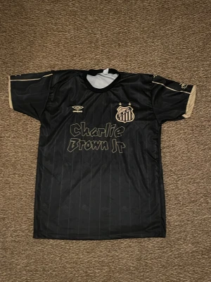 Svart Santos Neymar Jr fotbollströja - Snygg svart Santos FC fotbollströja från Umbro med guldiga detaljer och tryck. Framsidan har klubbmärke, två stjärnor och Charlie Brown Jr-tryck, medan baksidan har Neymar Jr och nummer 10. Tunn, lätt polyester med diskreta ränder och korta ärmar.