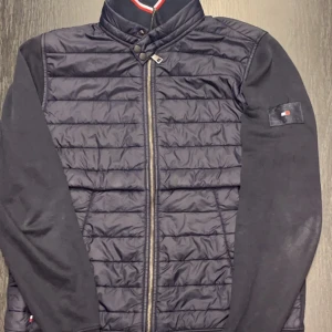 Marinblå jacka från Tommy Hilfiger - Snygg marinblå jacka från Tommy Hilfiger med quiltad framsida och dragkedja. Jackan har långa ärmar och en liten logotyp på ärmen. Tillverkad i en blandning av ekologisk bomull. Jackan har tappat färgen vid armarna, tommy hilfiger märket är lite slitet.