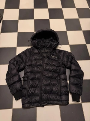 Svart pufferjacka från Peak Performance - Säljer en svart pufferjacka från Peak Performance med huva och dragkedja. Jackan har quiltad design, två sidofickor med dragkedja och ribbade muddar vid ärmsluten. Perfekt för kalla dagar och riktigt snygg till streetwear-stilen.