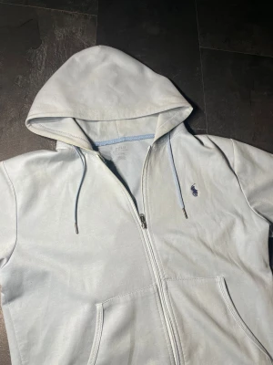 Ljusgrå hoodie från Polo Ralph Lauren - Snygg ljusgrå hoodie från Polo Ralph Lauren med dragkedja och huva. Tröjan har broderad mörkblå logga på bröstet, snörning i huvan och två praktiska fickor framtill. Tillverkad i mjuk bomull för skön komfort. Perfekt för chill dagar.