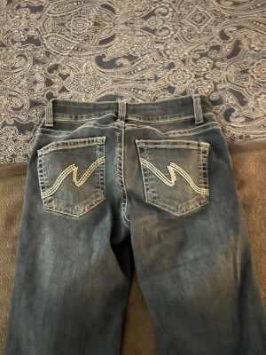 Blå bootcut jeans från Nelly - Snygga blå jeans från Nelly med bootcut-ben och klassisk femficksdesign. Jeansen har ljusa kontrastsömmar och coola broderade detaljer på bakfickorna. Materialet är jeans med en något mörk tvätt och dubbla knappar i midjan.