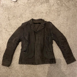 Svart parkajacka från Diesel - Svart parkajacka från Diesel med coola detaljer som stora fickor framtill, axelklaffar och knappar vid ärmslut. Jackan är tillverkad i bomull och har en klassisk krage. Perfekt för dig som gillar streetstyle och vill ha en snygg jacka med attityd.