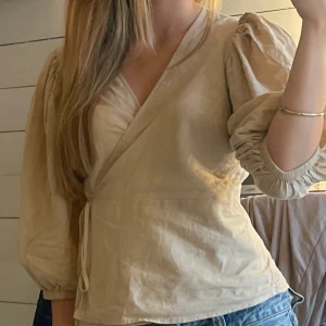 Beige omlottblus med puffärm - Säljer en beige omlottblus med puffiga trekvartsärmar och knyt i midjan. Blusen har v-ringad hals och är tillverkad i ett lätt bomullsmaterial som ger en skön känsla. Perfekt att styla med jeans för en chill look.
