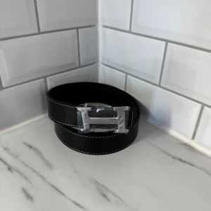 Svart bälte med H-spänne i silver - Snyggt svart bälte med ett stilrent H-format spänne i silverfärgad metall. Bältet har vit kontrastsöm längs kanterna och är tillverkat i präglat skinn som ger en lyxig känsla. Perfekt för att lyfta din outfit med en modern touch.