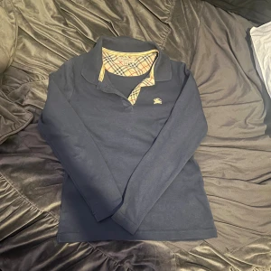 Mörkblå pikétröja från Burberry BARN - Snygg mörkblå pikétröja från Burberry med klassisk krage och det ikoniska rutiga mönstret på insidan av kragen. Tröjan har guldigt broderat Burberry-logo på bröstet och är långärmad. Perfekt för dig som vill ha en stilren och tidlös look. BARN SIZE I S