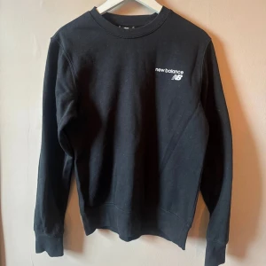 Svart sweatshirt från New Balance - Svart sweatshirt från New Balance med vit logga på bröstet. Tröjan har rund hals, ribbade muddar och är tillverkad i mjuk bomull. Perfekt för chill dagar eller när du vill ha en clean och enkel look. S