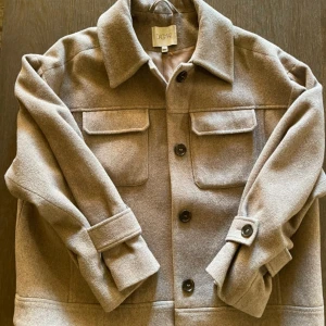 Beige jacka från Dagmar - Snygg beige jacka från Dagmar med klassisk krage, stora bröstfickor och knappar framtill. Jackan har långa ärmar med knappdetalj vid mudden och är tillverkad i mjuk ull. Perfekt för lager-på-lager och chill stil.