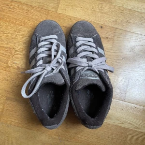 Adidas Campus sneakers brun mocka 38⅔ - Säljer ett par Adidas Campus sneakers i mörkbrun mocka med klassiska ränder på sidorna. Skorna har rund tå, platt sula och snörning med beige skosnören, finns mörkbruna också. Ikonisk Adidas-logga på plösen och insidan. Använda max 10 ggr. 