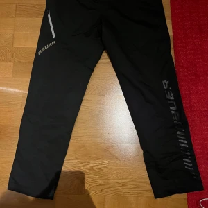 Svarta täckbyxor från Bauer - Svarta täckbyxor från Bauer med snyggt tryck längs benet och en praktisk dragkedja vid fickan. Byxorna har elastisk midja och är gjorda i slitstarkt syntetmaterial, perfekta för kalla dagar och sportaktiviteter. M och S size