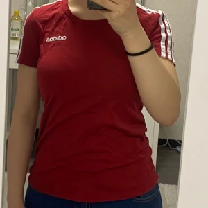 Röd Adidas t-shirt med vita ränder - Snygg röd t-shirt från Adidas med klassiska vita ränder längs axlarna och ärmarna. T-shirten har rund hals och korta ärmar, samt Adidas-logga broderad på bröstet. Perfekt för dig som gillar sportig och stilren look.