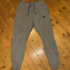Grå mjukisbyxor från Nike (Nike tech fleece), storlek S - Snygga grå mjukisbyxor från Nike i storlek S. Byxorna har svarta snören i midjan, ribbade muddar vid bensluten och praktiska fickor, varav en med dragkedja bak. Materialet är mjukt och skönt, perfekt för chill eller träning. Nike-logga i svart på framsidan.
