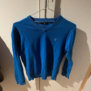 Blå v-ringad tröja från GANT - Blå v-ringad tröja från GANT med diskret logga broderad på bröstet. Tröjan har långa ärmar och ribbade muddar vid hals, ärmslut och nederkant. Perfekt för dig som gillar klassisk och stilren design.