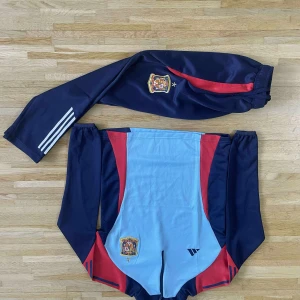 Spanska träningsbyxor Adidas - Säljer ett par mörkblå träningsbyxor från Adidas med Spaniens landslagsmärke och röda detaljer. Byxorna har elastisk midja med snörning och vita Adidas-ränder på benet. Materialet är polyester, perfekt för sport och fotboll.