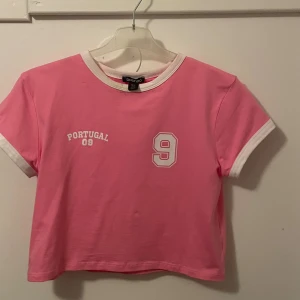 Rosa croppad t-shirt Portugal 09 Boohoo - Snygg rosa croppad t-shirt från Boohoo med vit text 'Portugal 09' och stort nummer 9 på både fram- och baksidan. T-shirten har vita detaljer vid hals och ärmslut, korta ärmar och sportig vibe. Perfekt för dig som gillar statement-plagg. 