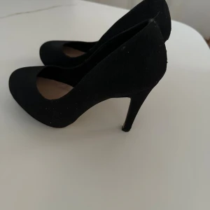 Svarta pumps från Bianco med klack - Snygga svarta pumps från Bianco med elegant hög klack och rund tå. Skorna har en stilren design och är klädda i ett tygmaterial som ger en mjuk känsla. Perfekta för dig som gillar klassisk och tidlös stil med en modern touch.