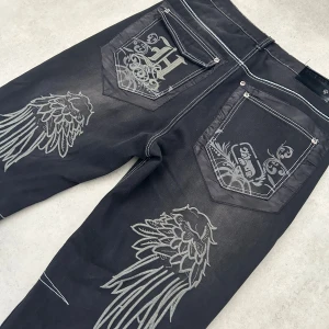Svarta jeans med tribal-vingar - Unika svarta jeans med grå tribal- och vingmönster på bakfickor och ben. Kontrasterande vita sömmar och klassisk femficksdesign. Snygga detaljer med broderade motiv som ger en edgy vibe. Perfekt för dig som vill sticka ut med din stil.