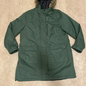 Grön parkajacka med fuskpäls - Snygg mörkgrön parkajacka med huva och fuskpäls. Jackan har långa ärmar, stora fickor med knappar och en praktisk bröstficka med dragkedja. Perfekt för kalla dagar och har en stilren look som funkar till många outfits. Kostar 99:-kr