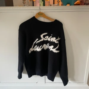 Svart stickad Saint Laurent tröja - Svart stickad tröja från Saint Laurent med vit, fluffig logga framtill i cool graffiti-stil. Tröjan har rund halsringning, långa ärmar och ribbade muddar. Perfekt för dig som vill ha en statement piece med lyxig känsla.