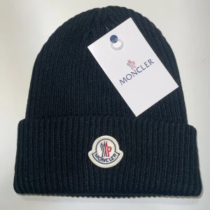 Svart ribbstickad mössa från Moncler - Svart ribbstickad mössa från Moncler med klassisk uppvikt kant och broderad logotyp framtill. Tillverkad i mjukt ullmaterial som håller dig varm under kyliga dagar. Perfekt accessoar för en stilren och sportig look.