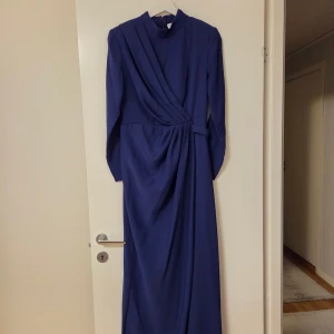 Marinblå långklänning med drapering - Snygg marinblå långklänning med lång ärm och hög hals. Klänningen har cool drapering framtill och markerad midja som ger en elegant siluett. Perfekt för dig som vill sticka ut med stilren look och klassiska detaljer.