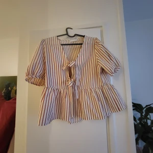 Randig blus Carin Wester - Supersöt blus med röda och vita ränder, v-ringning och två knyt framtill. Blusen har korta puffärmar med resår och volang nedtill som ger en flowy känsla. Perfekt för dig som gillar en trendig och luftig look.