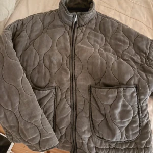 Grå quiltad jacka från Zara - Quiltad jacka från Zara i grå manchester med vågigt mönster. Jackan har två stora fickor framtill, hög krage och dragkedja. Den har en loose passform och är perfekt för dig som gillar chill och trendig stil.