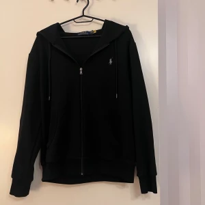 Polo Ralph Lauren zip up - Svart hoodie från Polo Ralph Lauren med broderad logga på bröstet. Tröjan har huva med snörning, dragkedja framtill och två fickor. Tillverkad i mjuk bomull och har långa ärmar med ribbade muddar. Perfekt för chill dagar och streetwear.