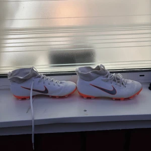 Vita Nike fotbollsskor med orange sula - Säljer ett par vita Nike fotbollsskor med orange och vit dobbsula. Skorna har snörning, högt skaft och en snygg svart Nike-logga på sidan. Ovandelen är i syntetmaterial och sulan är designad för bra grepp på planen. Perfekta för dig som vill sticka ut på matchen!