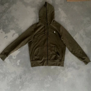 Olivgrön hoodie från Polo Ralph Lauren - Snygg olivgrön hoodie från Polo Ralph Lauren med klassisk vit broderad logga på bröstet. Tröjan har huva med snörning, hel dragkedja och en stor magficka framtill. Tillverkad i mjuk bomull, perfekt för chill dagar. Passformen är normal.